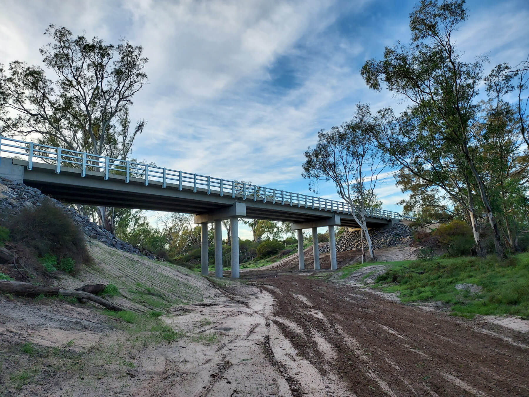 Photo looking from a dry river bed up to a large new concrete bridge. There are large gum trees on either side of the bridge and blue sky.