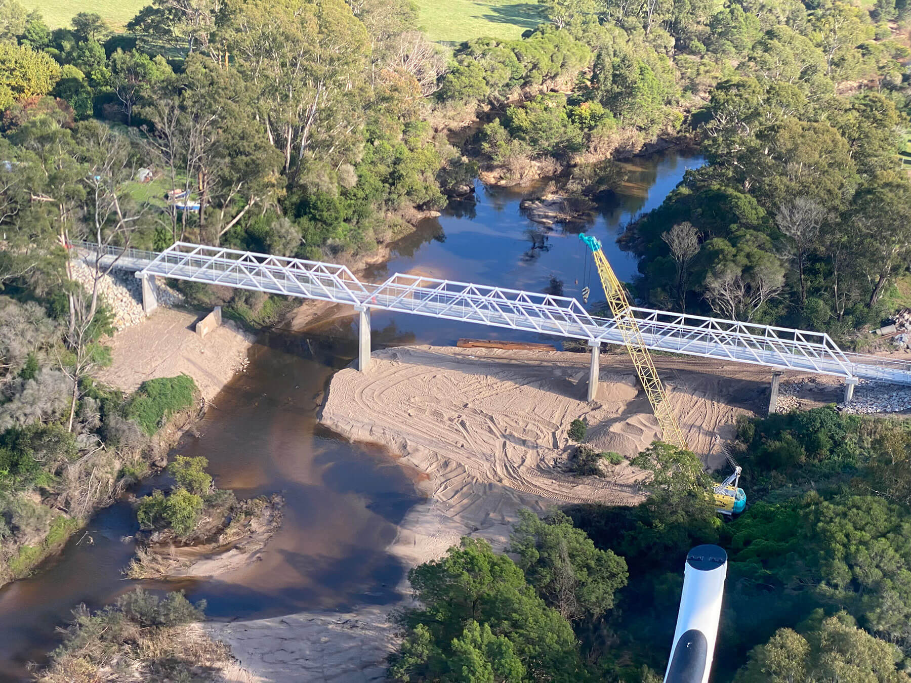 Aerial photo looking down at a new bridge being constructed over a river. There are two large cranes on the banks of the river, the bridge frame is silver and taking shape, and the site is in the countryside and surrounded by trees and mountains.