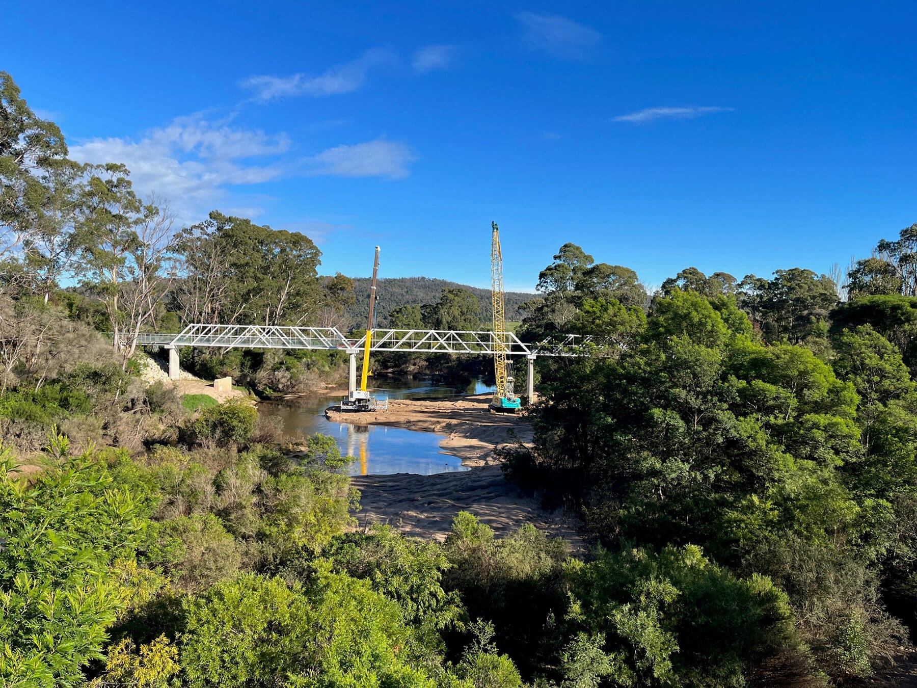 Chris-obrien-GENOA-PED-BRIDGE-3 Photo looking down a river at a new bridge being constructed over it. There are two large cranes on the banks of the river, the bridge frame is silver and taking shape, and the site is in the countryside and surrounded by trees and mountains.