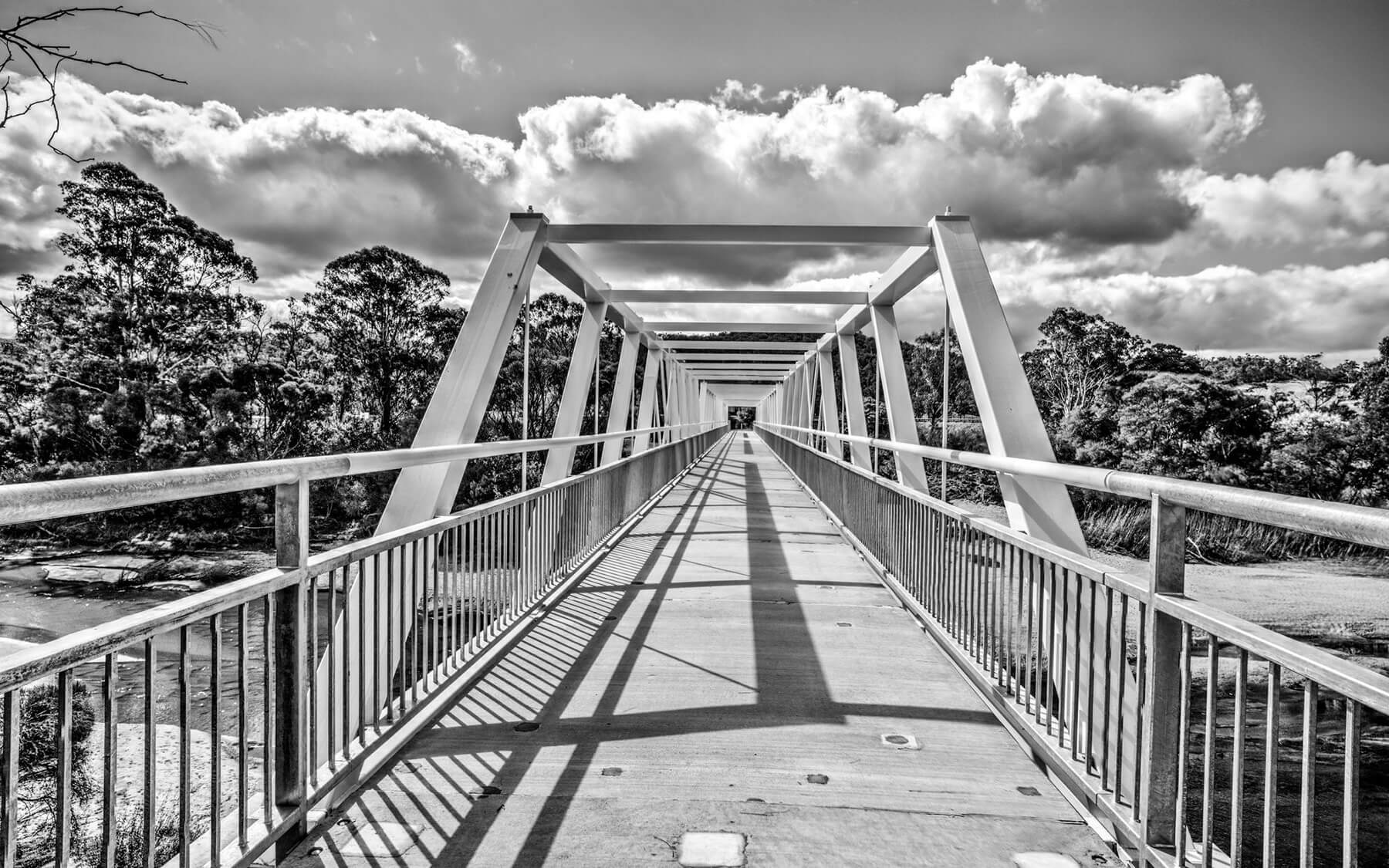 Black and white photo of a new footbridge, from standing at one side and looking across the bridge. The bridge has a concrete base with shiny silver handrails and sides, and overhead for the length of the bridge is a square metal frame with angles, that creates a visual effect as you enter the bridge and walk towards it. The angled overhead frame is designed to reflect the historical sturcture ihe new bridge replaces.
