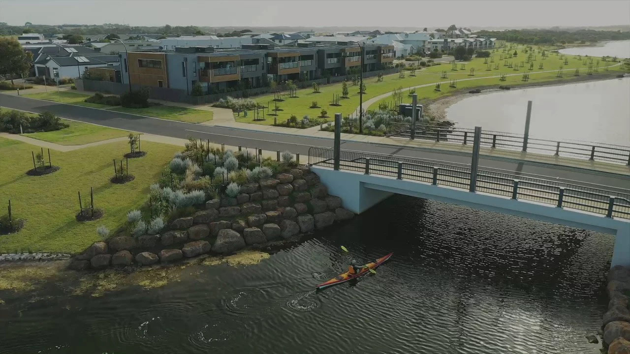 Aerial photo of a new bridge running over a waterway. There is lots of modern new housing, wide open lawns and newly planted trees long the waterway, and a person canoeing under the bridge.
