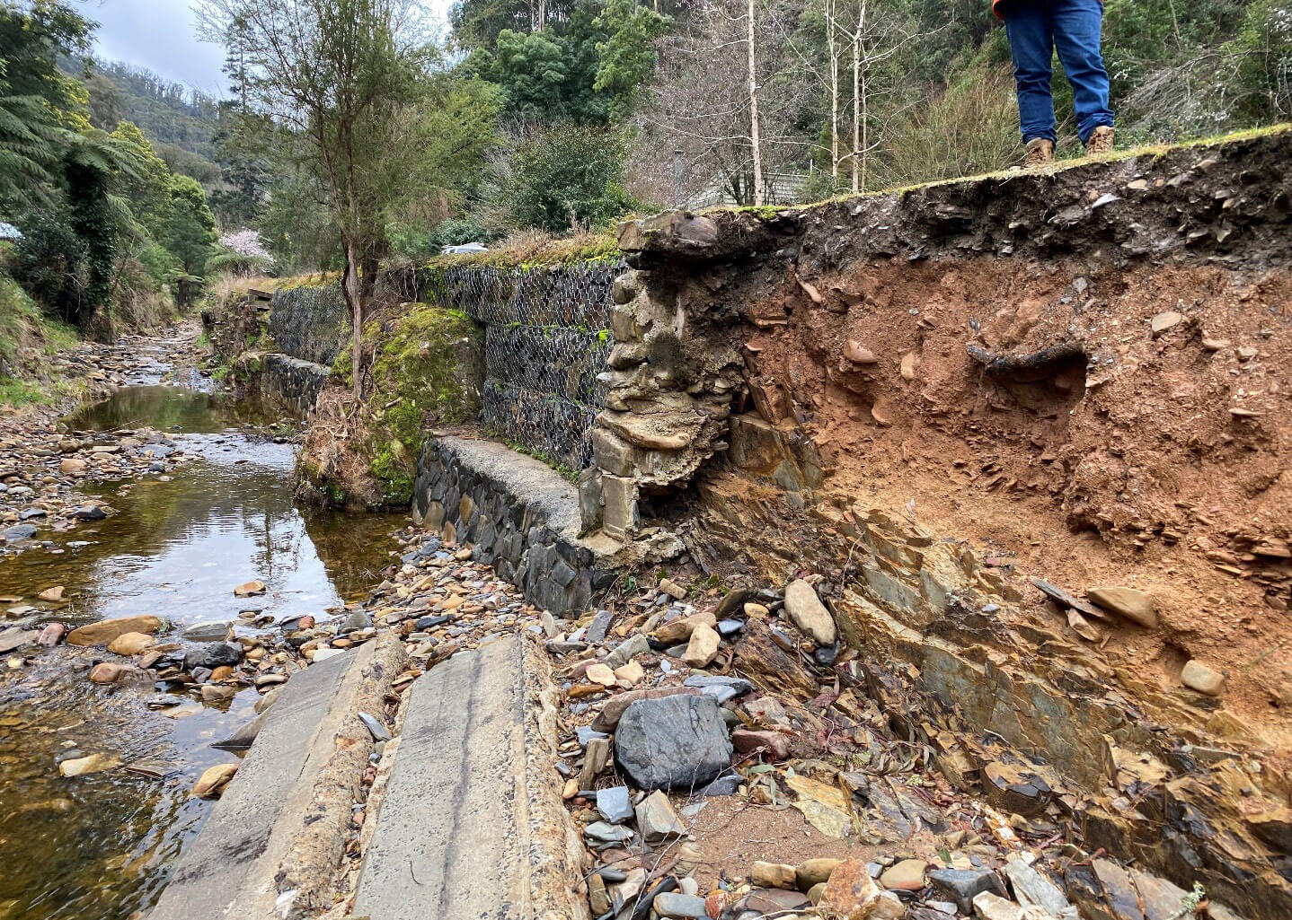 Close up photo of where a road has washed away during flooding of a small creek. Lots of rocks and pieces of concrete in the foreground, some chicken wire and in the distance the creek runs away into the mountains.