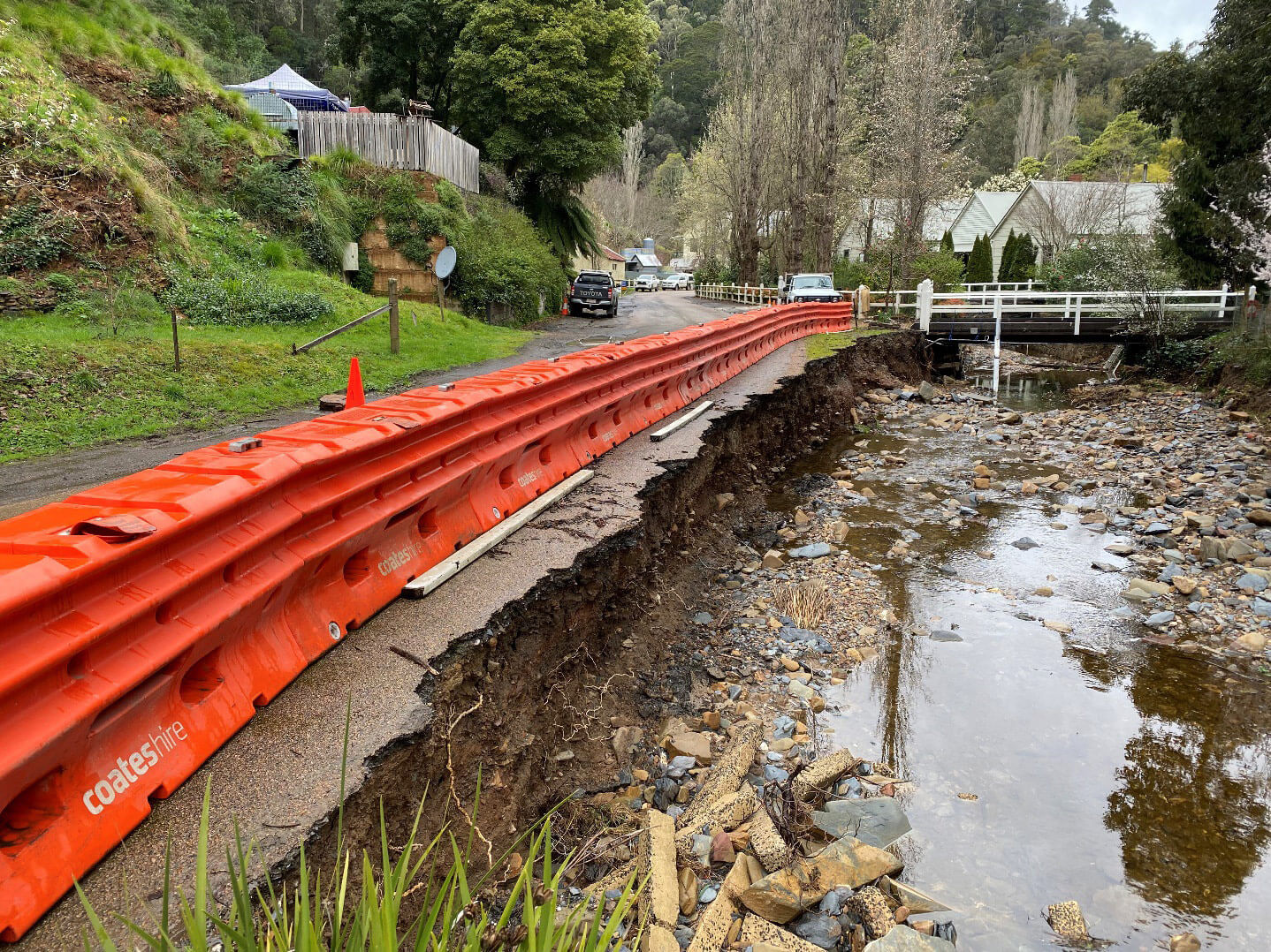 Photo of the side of a road washed away by flooding. The waterway is now not flooding but is filled with rocks and things left behind by the flood. There is a historical house on the right and other historic buildings dotted in the hillside around this. Orange safety barriers run along the edge of the road where it has washed away.