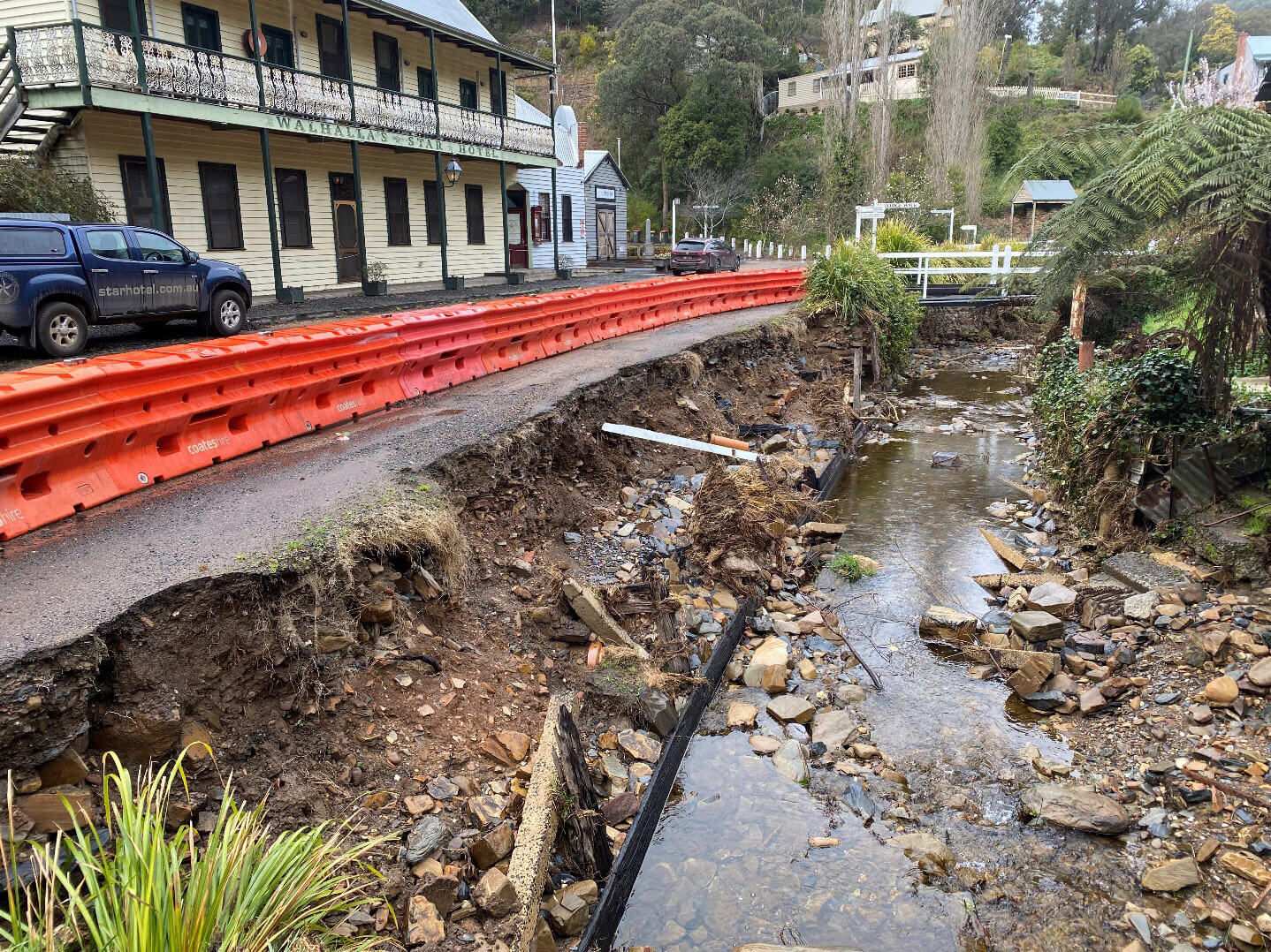 Photo of the side of a road washed away by flooding. The waterway is now not flooding but is filled with rocks and things left behind by the flood. There is a historical two story hotel on the left and other historic buildings dotted in the hillside around this. Orange safety barriers run along the edge of the road where it has washed away.