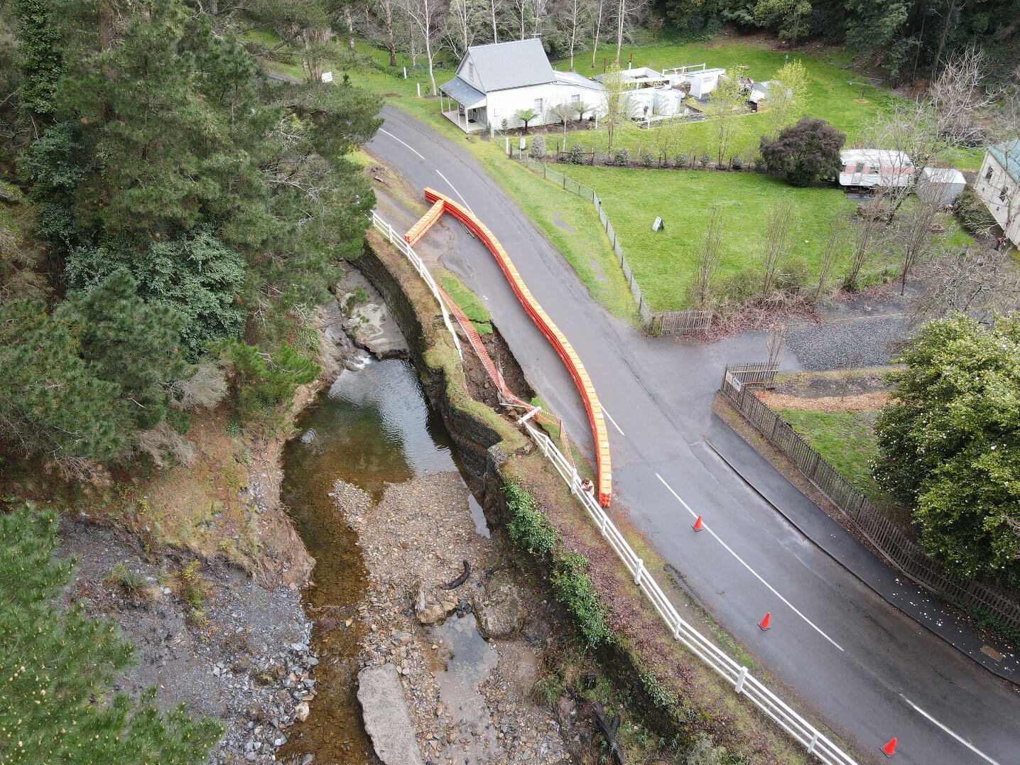 Chris-obrien-WALHALLA-FLOOD-RECOVERY-4 Aerial photo of a country road running alongside a small waterway. Flooding has caused the road to be washed away in some sections and these are blocked off to the public with orange safety barriers. The flooding is gone. There is a small white country house next to the road and lots of trees.
