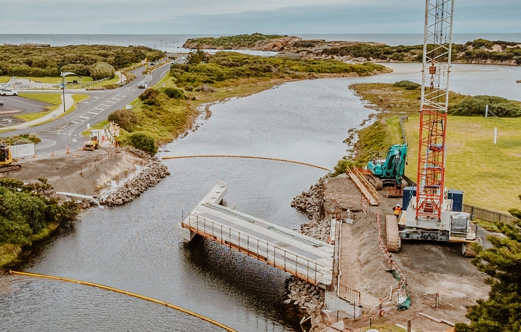 Chris-obrien-page-hydrology Aerial photo of a new bridge being built over a river that runs into the ocean. The bridge is half way over the water, and there is a large crane on the right side.