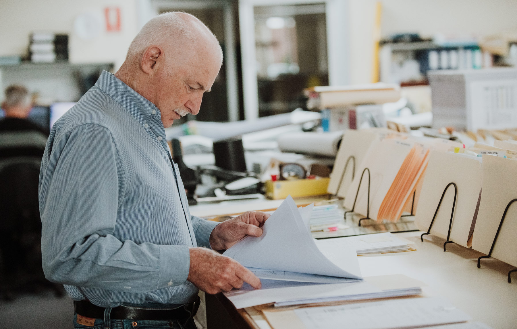 Chris-obrien-proof-engineering Photo of an older man standing, studying paperwork in an office. The main is wearing a light grey shirt with long sleeves and is surrounded by cream manilla folders and filing cabinets.