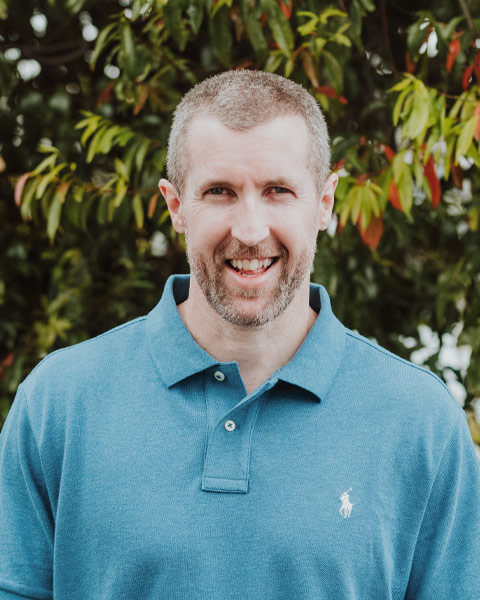 Chris-obrien-team-lukemilstein Photo of Luke Milstein wearing a teal blue polo shirt standing infront of green trees.