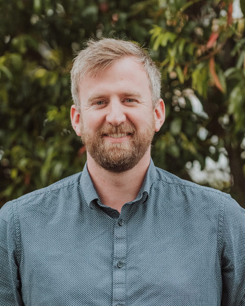 Chris-obrien-team-willhowden Photo of Will Howden wearing a blue/grey shirt standing infront of green trees.