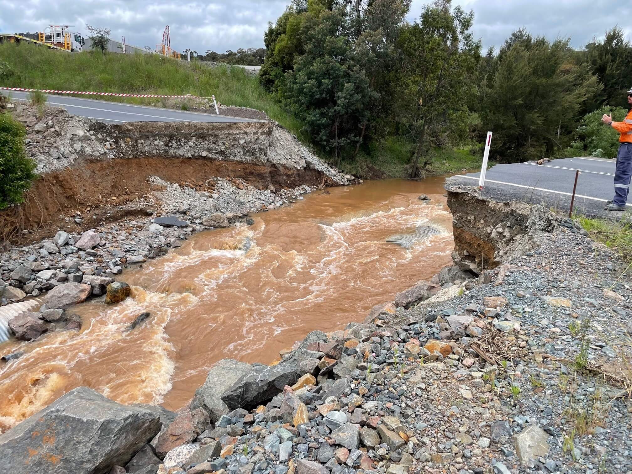 Photo of where a bridge used to be, it has been washed away. There is a big gap where the bridge used to be, like the road has collapsed into the river. Muddy brown water is flowing fast in the river, there are rocks and stones in the gap. You can see both sides of the road on the left and right side of the photo.