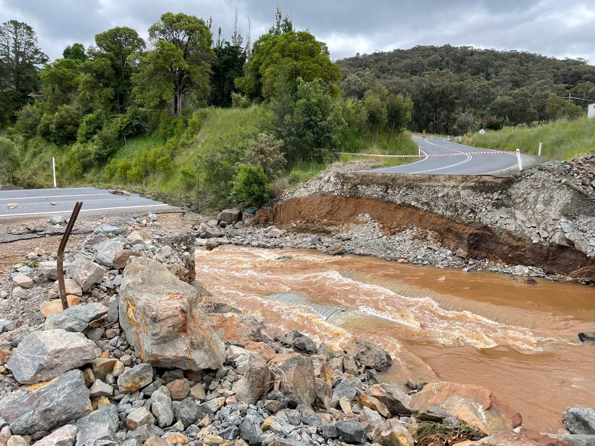 Photo of where a bridge used to be, it has been washed away. On the right the country road winds out of a hill and there is a big gap where the bridge used to be, like the road has collapsed into the river. Muddy brown water is flowing fast in the river, there are rocks and stones in the gap and on the left is the other side of the road.