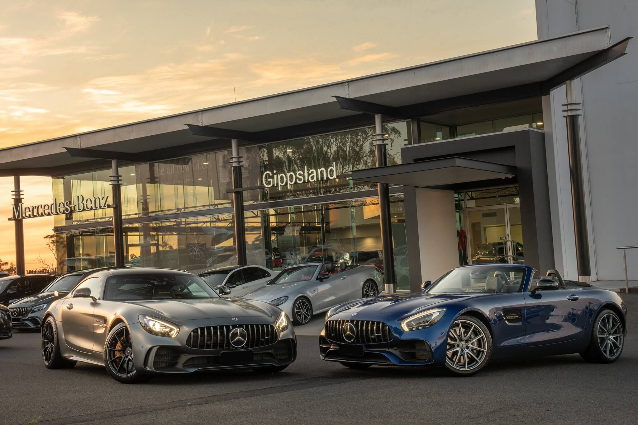Photo of a car dealership glass showroom for Mercedes-Benz, and a grey and a blue Mercedes sports cars parked infront of the building.