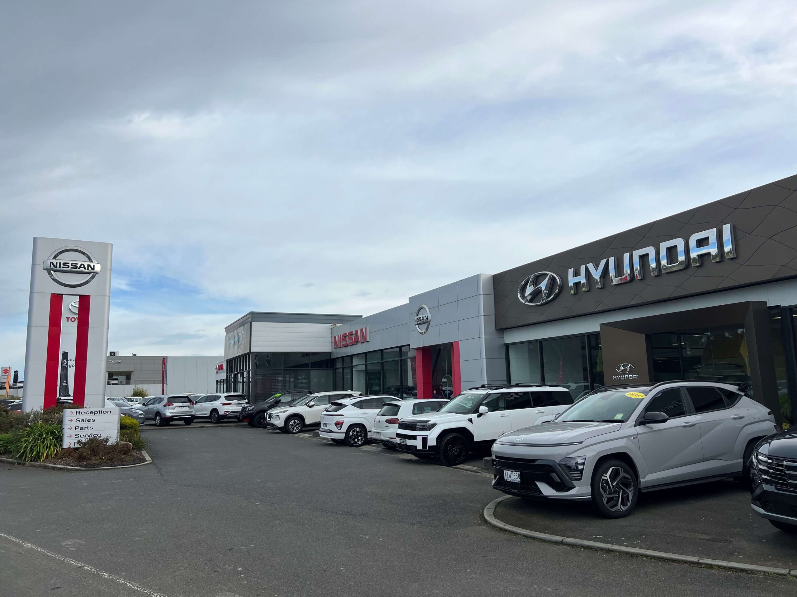 Photo of a car dealership showroom with large branding signs for Nissan and Hyundai on the buildings, and cars parked infront of the buildings.