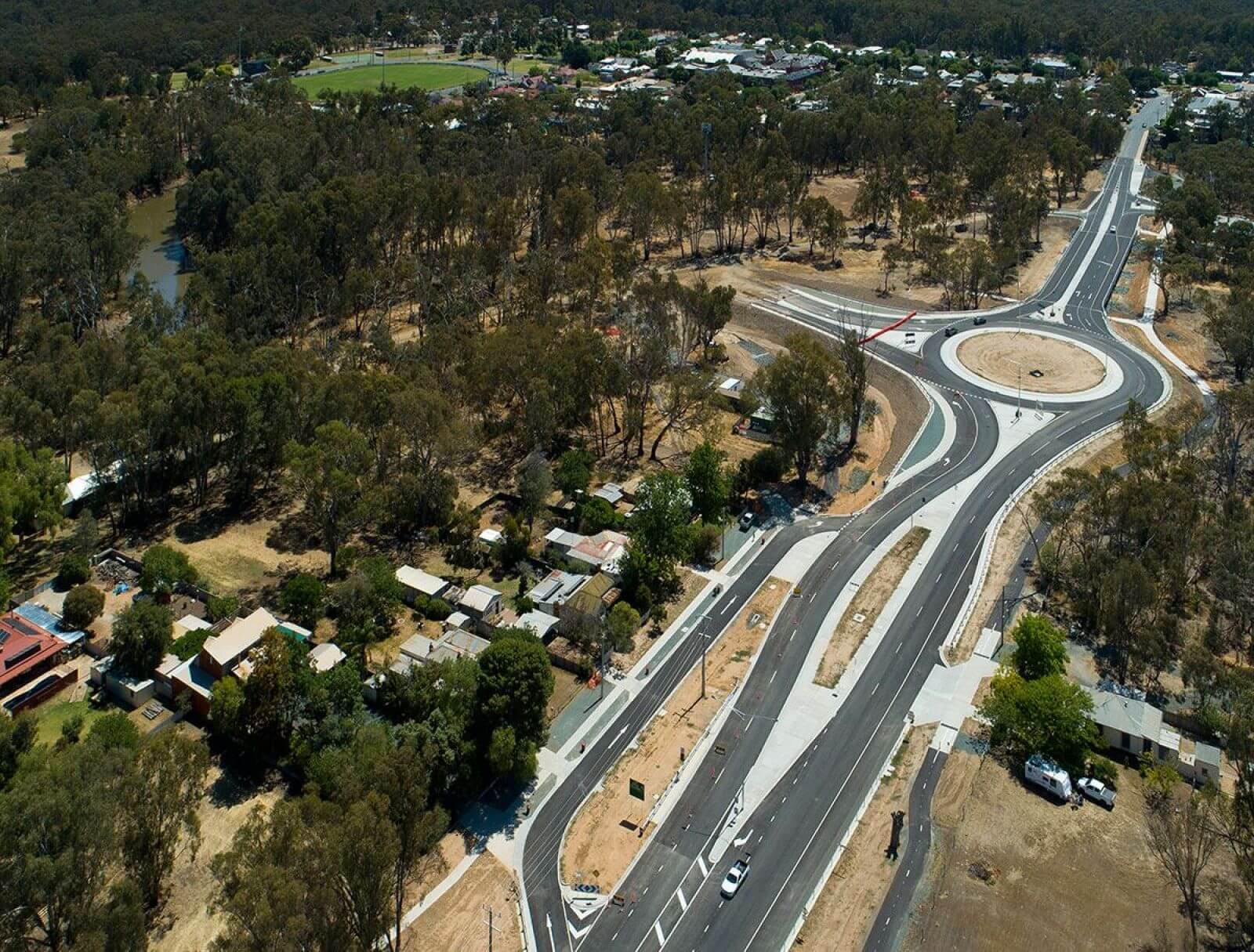 Aerial photo of a new highway with large roundabout. The landscape is dry countryside and green trees around it. It is a country area and there are houses and a sports oval in view.