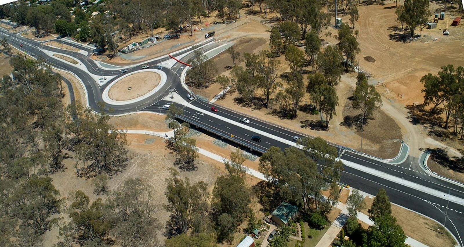 Aerial photo of a new highway with large roundabout. The landscape is dry countryside and green trees around it. There are a few houses around.