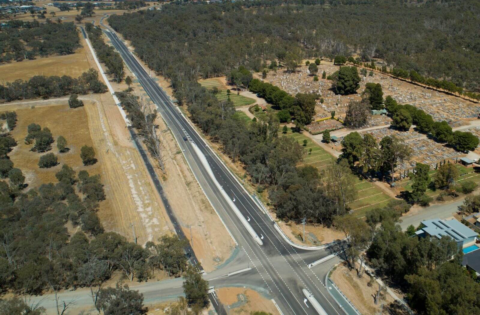 Aerial photo of a new highway, with dry countryside and green trees around it. On the left is a cemetery in the background.