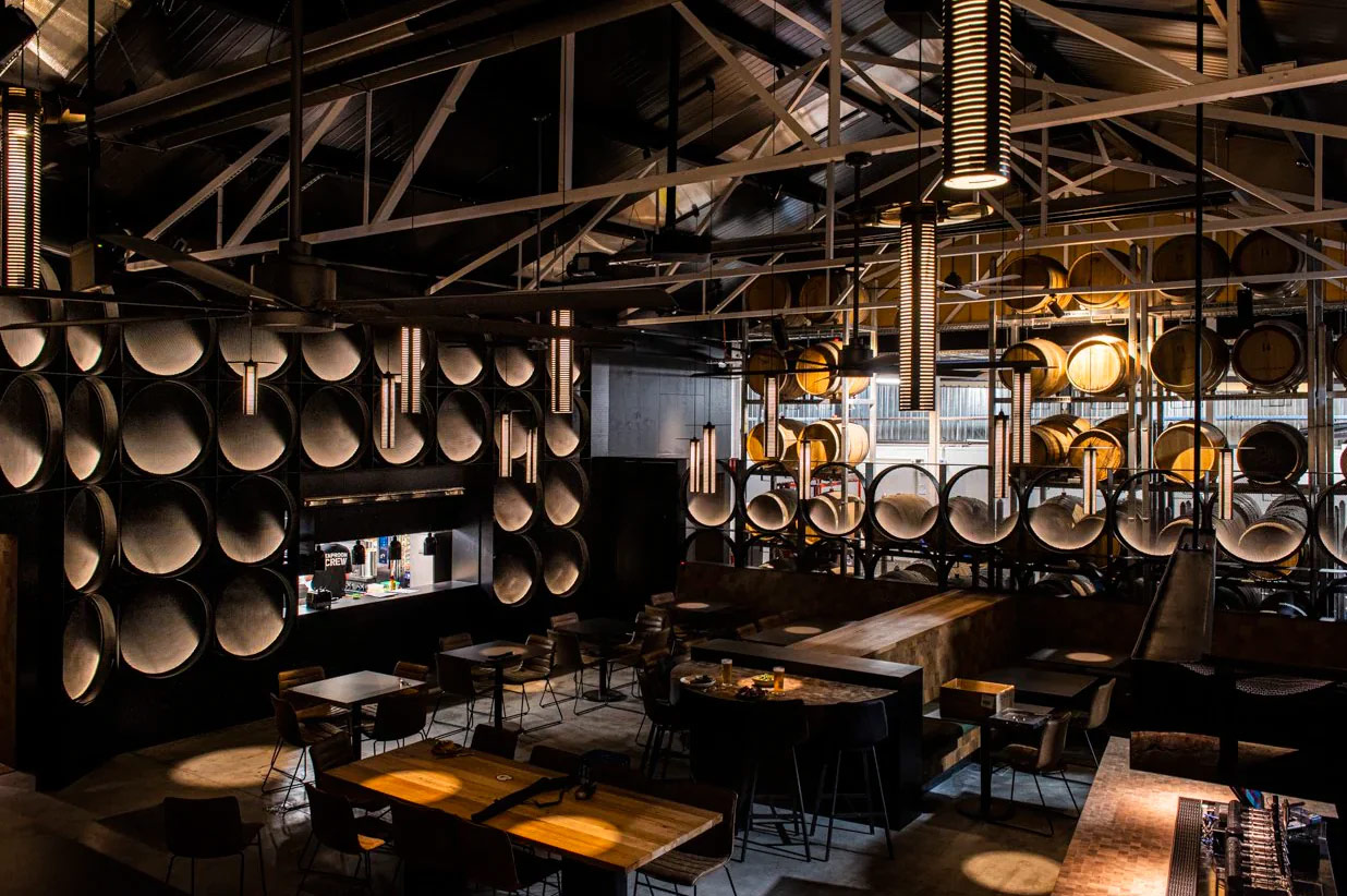 chris-obrien-deeds-brewery-1 Photo of the interior of a modern brewery with a long bar. The bar has golden lighting built into it in a circle pattern and overall the lighting is dark and moody. Overhead are many silver kegs hanging in a pattern from the roof and the space has a dramatic, stylish, modern look and feel.
