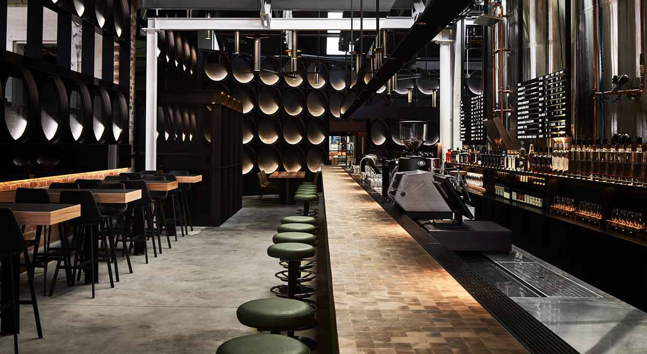 Photo of the interior of a modern brewery with a long bar. The bar has golden lighting built into it in a circle pattern and overall the lighting is dark and moody. Overhead are many silver kegs hanging in a pattern from the roof and the space has a dramatic, stylish, modern look and feel.