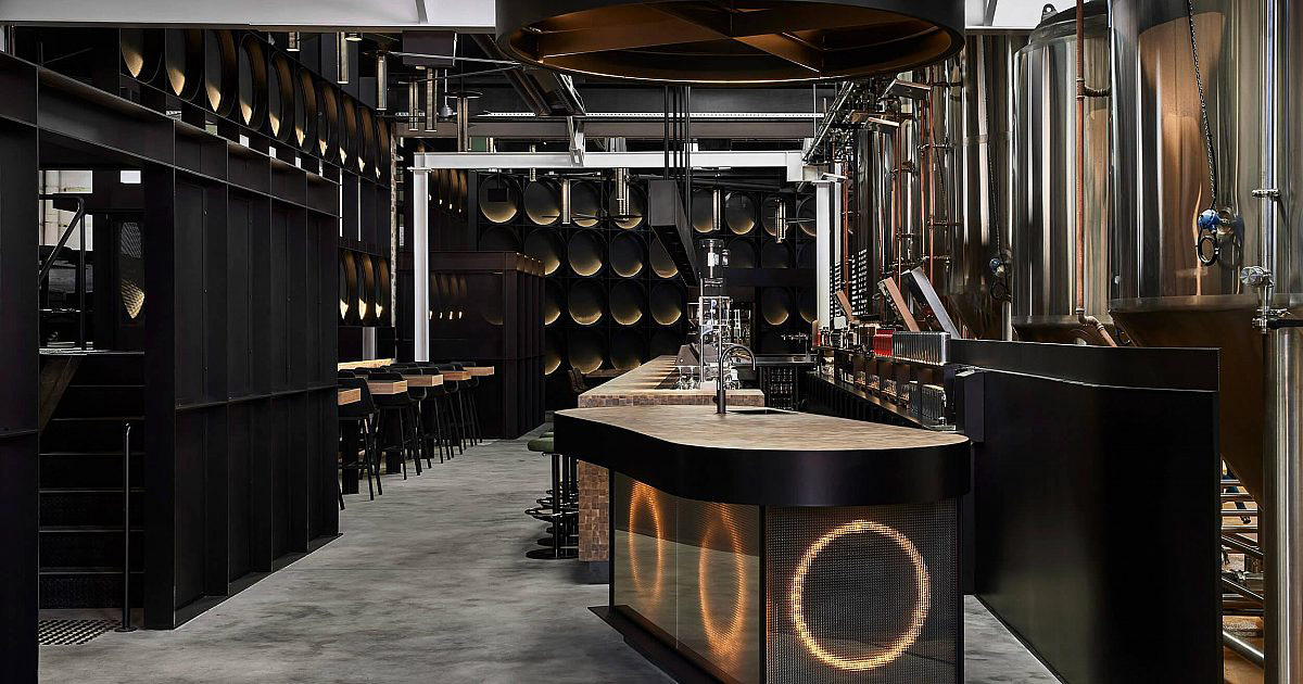 Photo of the interior of a modern brewery. The bar has golden lighting built into it in a circle pattern and overall the lighting is dark and moody. Overhead are many silver kegs hanging in a pattern from the roof and the space has a dramatic, stylish, modern look and feel.