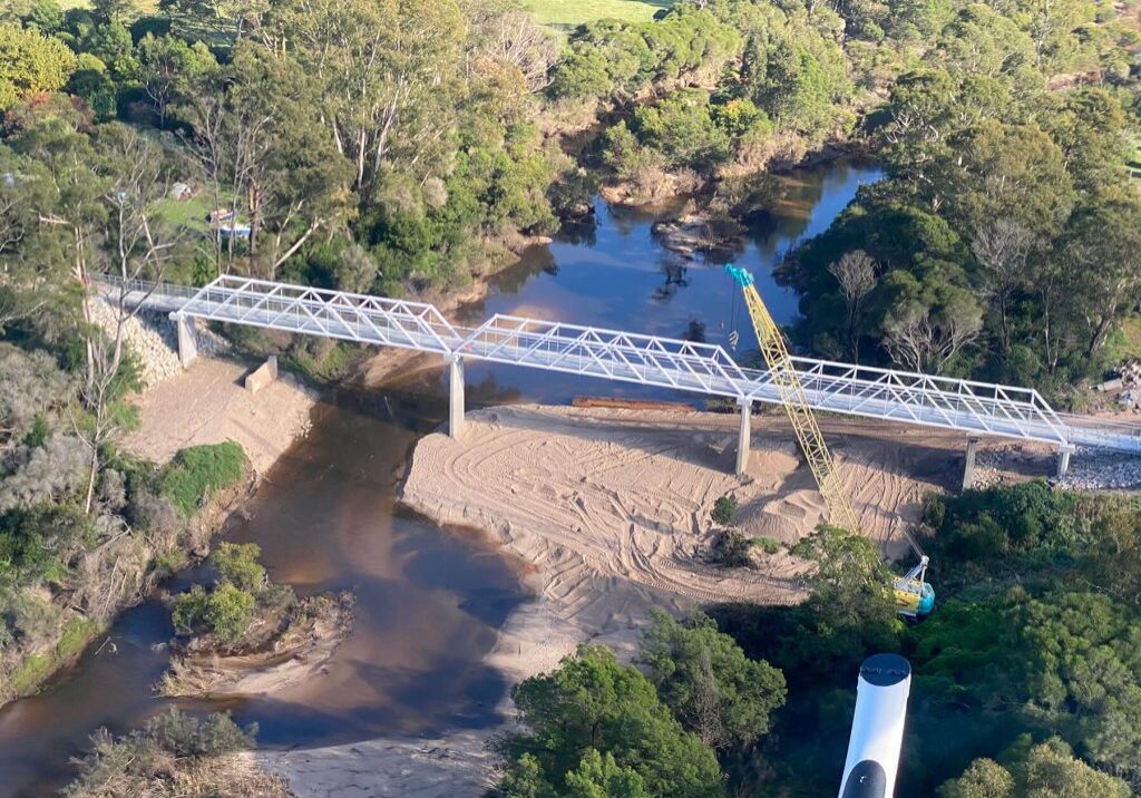 Aerial photo looking down at a new bridge being constructed over a river. There are two large cranes on the banks of the river, the bridge frame is silver and taking shape, and the site is in the countryside and surrounded by trees and mountains.