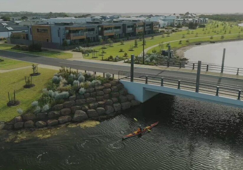 Aerial photo of a new bridge running over a waterway. There is lots of modern new housing, wide open lawns and newly planted trees long the waterway, and a person canoeing under the bridge.