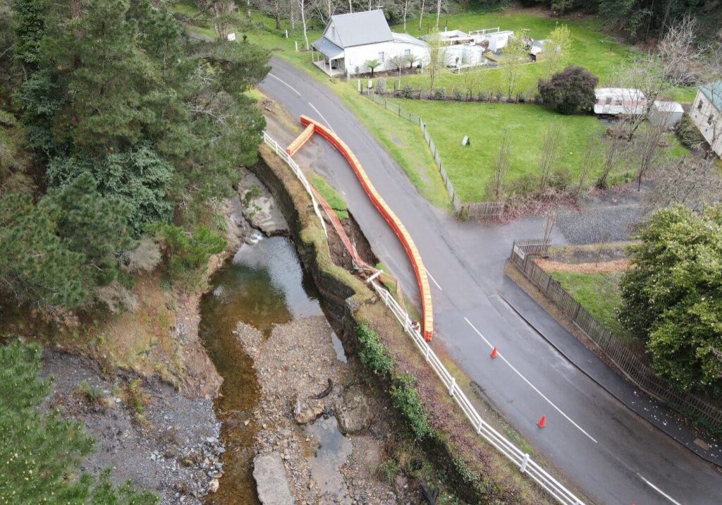 Aerial photo of a country road running alongside a small waterway. Flooding has caused the road to be washed away in some sections and these are blocked off to the public with orange safety barriers. The flooding is gone. There is a small white country house next to the road and lots of trees.