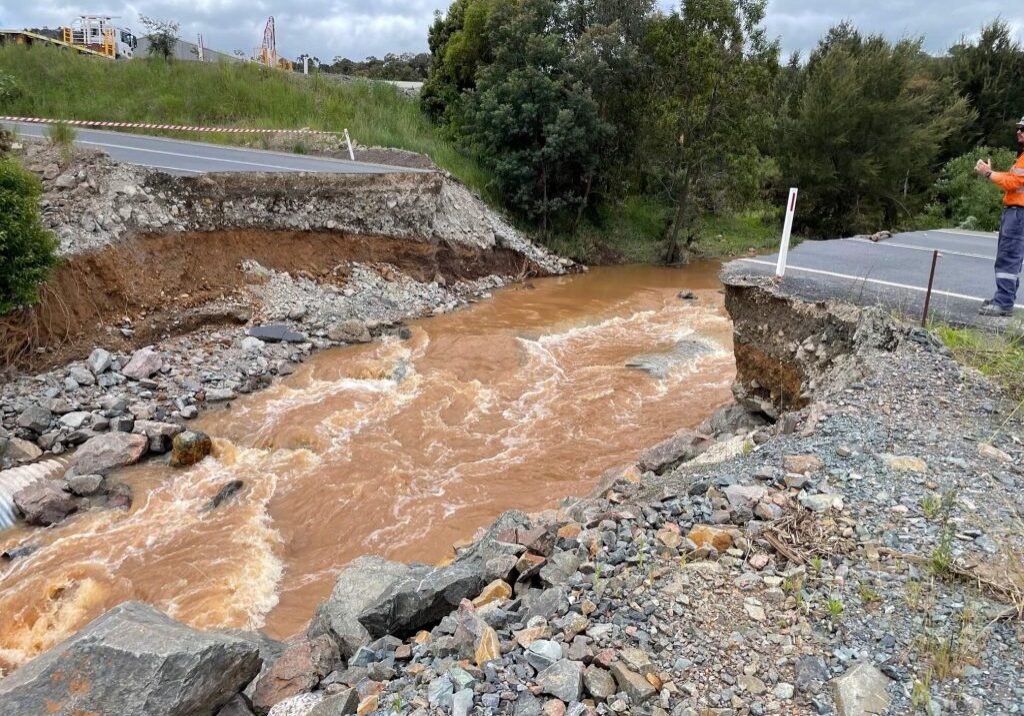Photo of where a bridge used to be, it has been washed away. There is a big gap where the bridge used to be, like the road has collapsed into the river. Muddy brown water is flowing fast in the river, there are rocks and stones in the gap. You can see both sides of the road on the left and right side of the photo.
