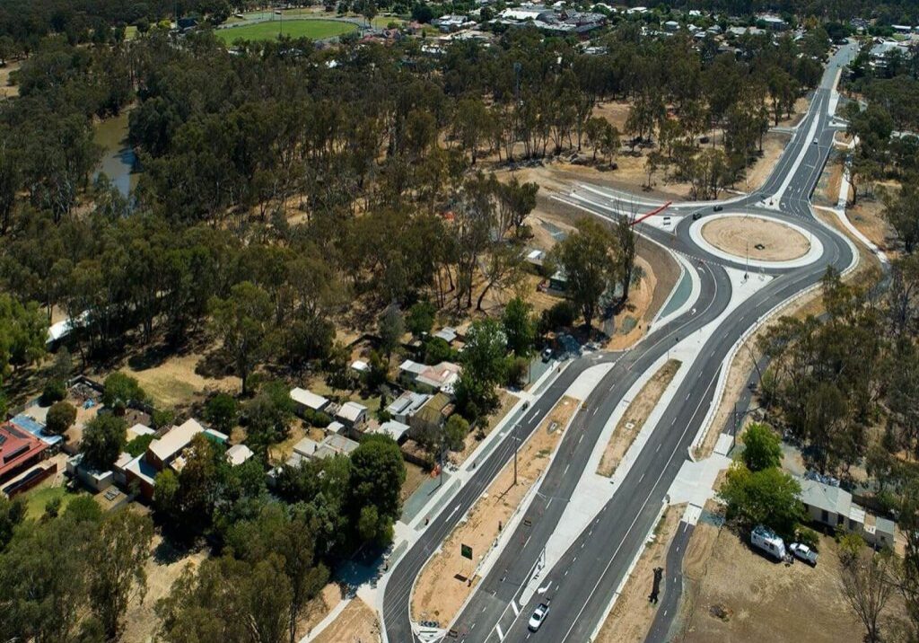 Aerial photo of a new highway with large roundabout. The landscape is dry countryside and green trees around it. It is a country area and there are houses and a sports oval in view.