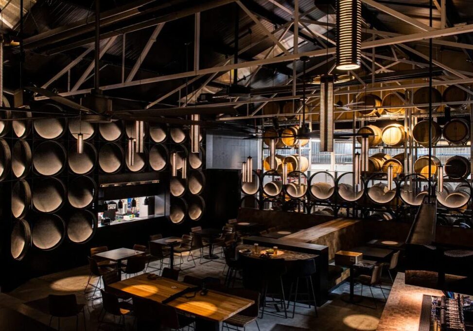 Photo of the interior of a modern brewery with a long bar. The bar has golden lighting built into it in a circle pattern and overall the lighting is dark and moody. Overhead are many silver kegs hanging in a pattern from the roof and the space has a dramatic, stylish, modern look and feel.